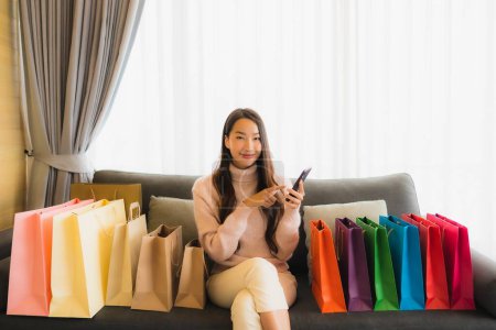 Photo for Portrait beautiful young asian woman using laptop or smart mobile cell phone for shopping online on sofa around shopping bag in living room interior - Royalty Free Image
