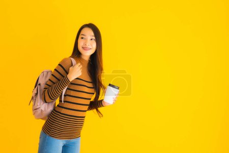 Téléchargez les photos : Portrait belle jeune femme asiatique avec sac à dos et tasse de café dans sa main sur fond jaune isolé - en image libre de droit