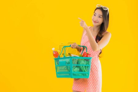 Téléchargez les photos : Portrait belle jeune femme asiatique avec épicerie dans le panier shopping de supermarché sur fond jaune isolé - en image libre de droit