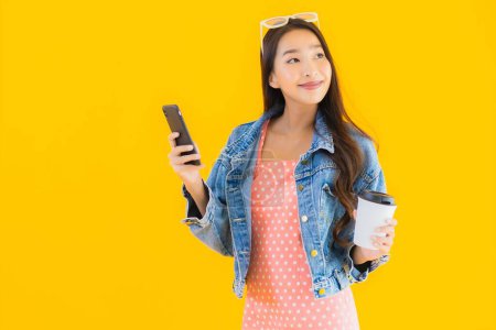 Téléchargez les photos : Portrait belle jeune femme asiatique avec tasse de café et téléphone portable intelligent sur fond jaune isolé - en image libre de droit