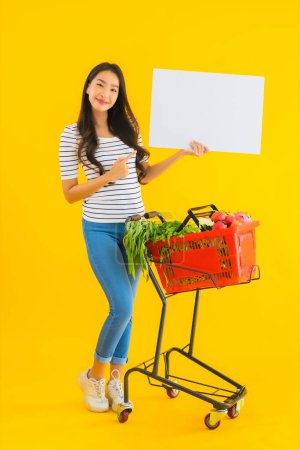 Foto de Retrato hermosa joven asiática mujer con carrito de la cesta de la compra y mostrar blanco tablero vacío sobre fondo aislado amarillo - Imagen libre de derechos