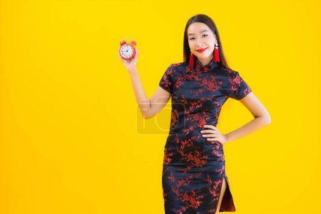 Téléchargez les photos : Portrait belle jeune femme asiatique porter robe chinoise montre horloge ou alarme sur fond jaune isolé - en image libre de droit