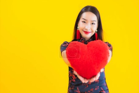 Téléchargez les photos : Portrait belle jeune femme asiatique porter robe chinoise montrer oreiller en forme de coeur sur fond jaune isolé - en image libre de droit