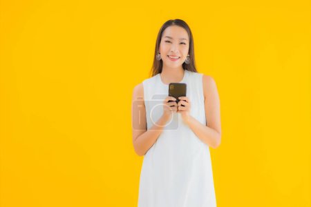 Téléchargez les photos : Portrait belle jeune femme asiatique avec téléphone portable intelligent sur fond jaune isolé - en image libre de droit