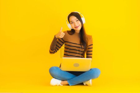 Photo for Beautiful portrait young asian woman sit on the floor with laptop and headphone on yellow isolated background - Royalty Free Image