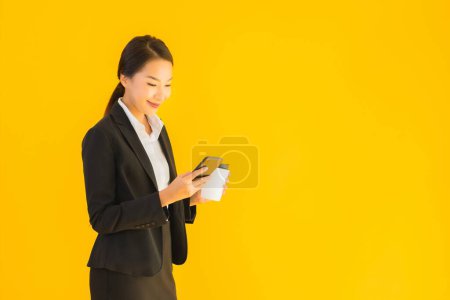 Téléchargez les photos : Portrait belle jeune femme asiatique d'affaires avec téléphone portable intelligent et tasse de café sur fond jaune isolé - en image libre de droit