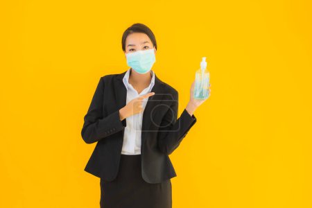 Téléchargez les photos : Portrait belle jeune entreprise asiatique femme porter masque utiliser gel d'alcool pour protéger de covid19 ou coronavirus sur fond jaune isolé - en image libre de droit