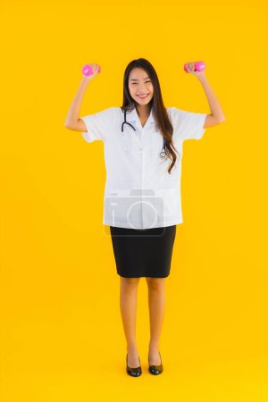 Téléchargez les photos : Portrait belle jeune asiatique médecin femme exercice avec haltère sur fond jaune isolé - en image libre de droit