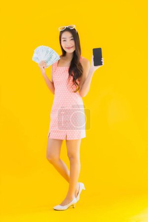 Foto de Retrato hermosa joven asiática mujer mostrar un montón de dinero en efectivo y dinero con el teléfono inteligente móvil en amarillo aislado fondo - Imagen libre de derechos