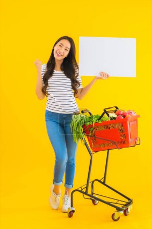 Photo for Portrait beautiful young asian woman with grocery basket cart and show white empty board on yellow isolated background - Royalty Free Image