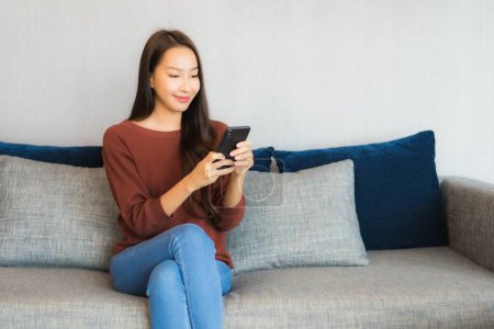 Téléchargez les photos : Portrait belle jeune femme asiatique utiliser téléphone intelligent sur canapé dans le salon intérieur - en image libre de droit
