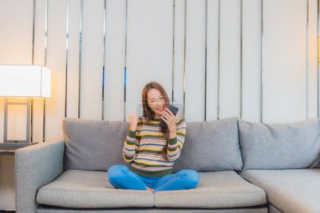Foto de Retrato hermosa joven asiático mujer uso inteligente móvil en sofá en sala de estar interior - Imagen libre de derechos