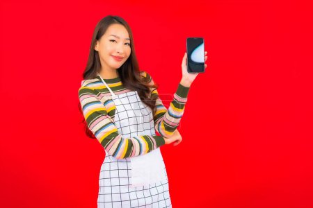 Foto de Retrato hermosa joven mujer asiática usar delantal con teléfono móvil inteligente en rojo aislado fondo - Imagen libre de derechos
