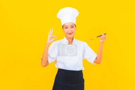 Téléchargez les photos : Portrait belle jeune femme asiatique porter chef ou cuisinier uniforme avec cuillère et fourchette sur fond jaune isolé - en image libre de droit