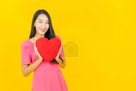 Téléchargez les photos : Portrait belle jeune femme asiatique sourire avec forme d'oreiller de coeur sur fond de couleur jaune - en image libre de droit