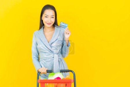 Téléchargez les photos : Portrait belle jeune femme asiatique sourire avec panier d'épicerie du supermarché sur fond de couleur jaune - en image libre de droit
