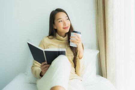 Téléchargez les photos : Jeune femme asiatique avec tasse de café et lire le livre sur le lit dans la chambre à coucher intérieure - en image libre de droit