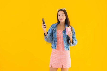 Téléchargez les photos : Portrait belle jeune femme asiatique profiter heureux avec téléphone pop-corn et regarder le film sur fond jaune isolé - en image libre de droit