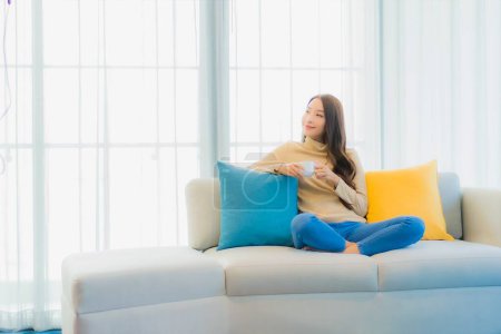 Foto de Retrato hermosa joven mujer asiática con taza de café en el sofá en el interior de la sala de estar - Imagen libre de derechos