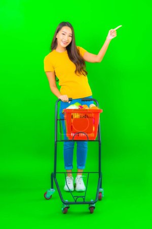 Foto de Retrato hermosa joven asiática mujer con cesta de comestibles y carrito de supermercado sobre fondo verde - Imagen libre de derechos