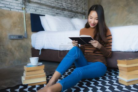 Photo for Young asian woman with coffee cup and read book on bed in bedroom interior - Royalty Free Image