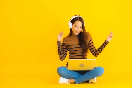 Téléchargez les photos : Beau portrait jeune femme asiatique assise sur le sol avec ordinateur portable et casque sur fond jaune isolé - en image libre de droit