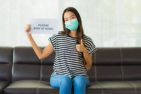 Photo for Portrait beautiful young with mask show paper stay home on sofa in livingroom for save people from coronavirus or covid19 - Royalty Free Image
