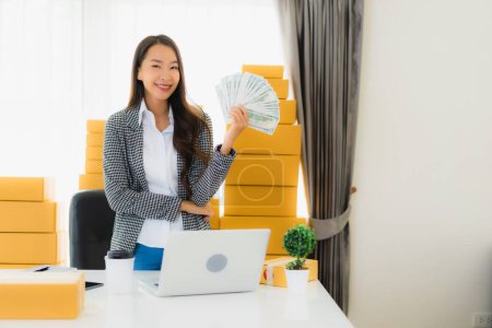 Foto de Retrato hermosa joven mujer asiática trabajo desde casa con portátil en efectivo y caja de cartón listo para el envío de compras de clientes en línea - Imagen libre de derechos