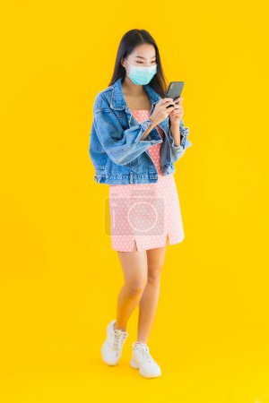 Foto de Retrato hermosa mujer asiática joven usar máscara para proteger covid19 o coronavirus utilizar el teléfono móvil inteligente sobre fondo aislado amarillo - Imagen libre de derechos
