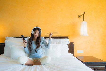 Foto de Retrato hermosa mujer asiática joven utilizando el teléfono celular móvil con auriculares para escuchar música en el interior del dormitorio - Imagen libre de derechos