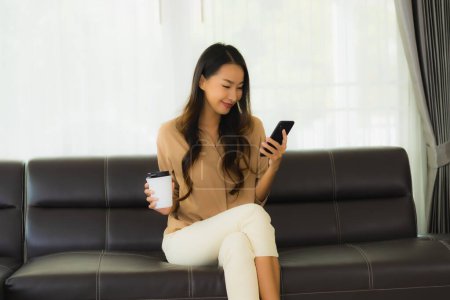 Foto de Retrato hermosa mujer asiática joven utilizar teléfono inteligente móvil o teléfono celular con taza de café en el interior del salón sofá - Imagen libre de derechos