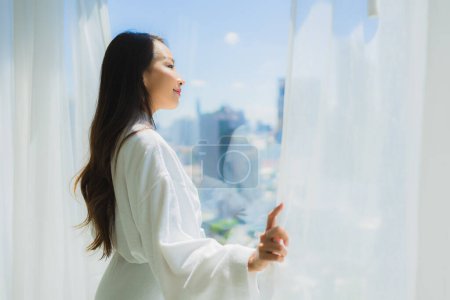 Téléchargez les photos : Portrait belle jeune femme asiatique ouverte fenêtre rideau et regarder à l'extérieur de la fenêtre pour la vue - en image libre de droit