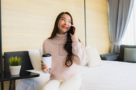 Téléchargez les photos : Portrait belle jeune femme asiatique avec tasse de café et téléphone mobile sur le lit dans la chambre à coucher intérieur - en image libre de droit