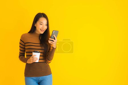 Foto de Hermosa joven asiática uso de teléfono móvil inteligente o teléfono celular con taza de café en muchas acciones sobre fondo aislado amarillo - Imagen libre de derechos