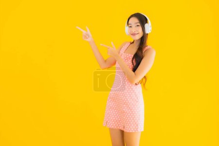 Téléchargez les photos : Portrait belle jeune femme asiatique écouter de la musique avec téléphone portable intelligent et casque sur fond jaune isolé - en image libre de droit