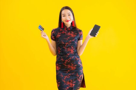 Foto de Retrato hermosa mujer asiática joven usar vestido chino con tarjeta de crédito y teléfono móvil inteligente sobre fondo aislado amarillo - Imagen libre de derechos