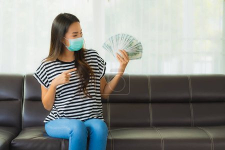 Foto de Retrato hermosa joven asiática mujer con máscara en sofá mostrar dinero o dinero en efectivo en casa - Imagen libre de derechos