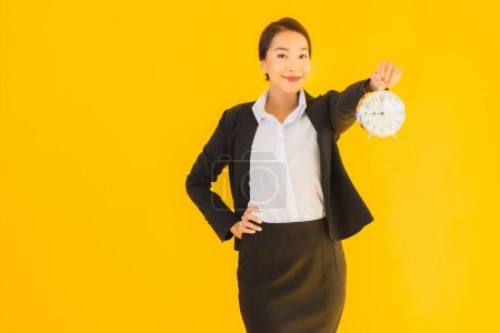 Téléchargez les photos : Portrait belle jeune femme asiatique montrer l'heure de l'horloge ou de l'alarme sur fond jaune isolé - en image libre de droit