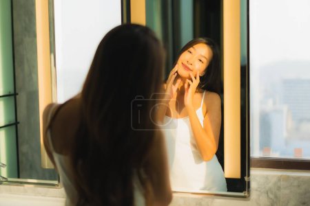 Téléchargez les photos : Portrait belle jeune femme asiatique vérifier son visage et le visage dans la salle de bain intérieure - en image libre de droit