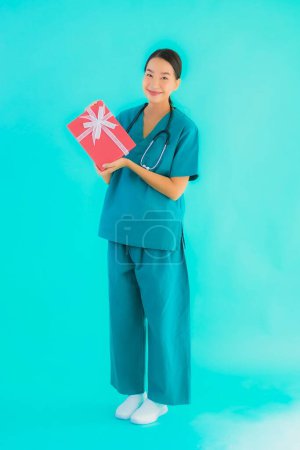 Téléchargez les photos : Portrait belle jeune asiatique médecin femme montrer boîte cadeau rouge sur fond bleu isolé - en image libre de droit