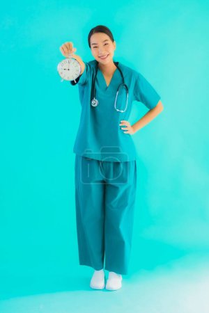 Foto de Portriat hermosa joven asiática médico mujer mostrar reloj o alarma en azul aislado fondo - Imagen libre de derechos