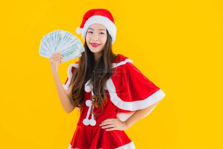 Foto de Retrato hermosa joven asiática mujer usar navidad ropa y sombrero con dinero en efectivo y dinero en amarillo aislado fondo - Imagen libre de derechos
