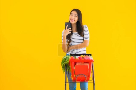 Foto de Retrato hermosa joven asiática mujer compras tienda de comestibles de supermercado y carro con teléfono móvil inteligente en amarillo aislado fondo - Imagen libre de derechos