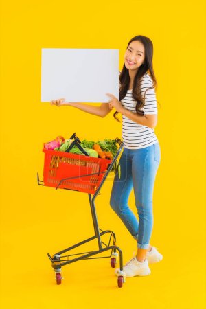 Téléchargez les photos : Portrait belle jeune femme asiatique avec panier d'épicerie chariot et montrer tableau blanc vide sur fond jaune isolé - en image libre de droit