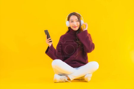Téléchargez les photos : Portrait belle jeune femme asiatique utiliser téléphone portable intelligent écouter de la musique sur fond jaune isolé - en image libre de droit