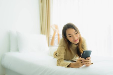 Téléchargez les photos : Jeune femme asiatique utilisant téléphone intelligent mobile sur le lit dans la chambre à coucher intérieur - en image libre de droit