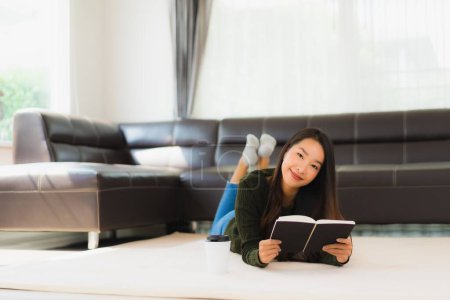 Foto de Retrato hermosa joven asiática mujer leer libro con taza de café y sofá en salón interior - Imagen libre de derechos