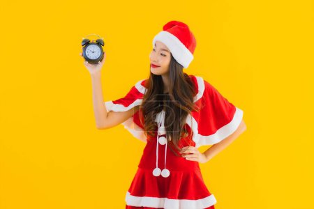 Téléchargez les photos : Portrait belle jeune femme asiatique porter des vêtements de Noël et chapeau montre horloge ou alarme sur fond isolé jaune - en image libre de droit