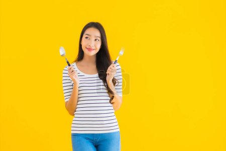 Téléchargez les photos : Portrait belle jeune femme asiatique montrer cuillère et fourchette prêt à manger sur fond jaune isolé - en image libre de droit