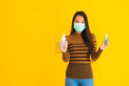 Téléchargez les photos : Portrait belle jeune femme asiatique avec masque vaporisateur d'alcool et bouteille de gel dans sa main pour protéger contre le coronavirus ou covid19 sur fond isolé jaune - en image libre de droit